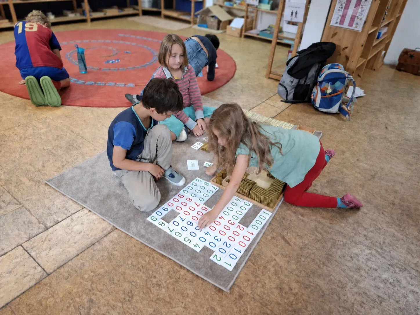Drei Kinder arbeiten mit Stellenwertkarten und goldenem Montessori-Perlenmaterial; im Hintergrund liegt eine Tausenderkette