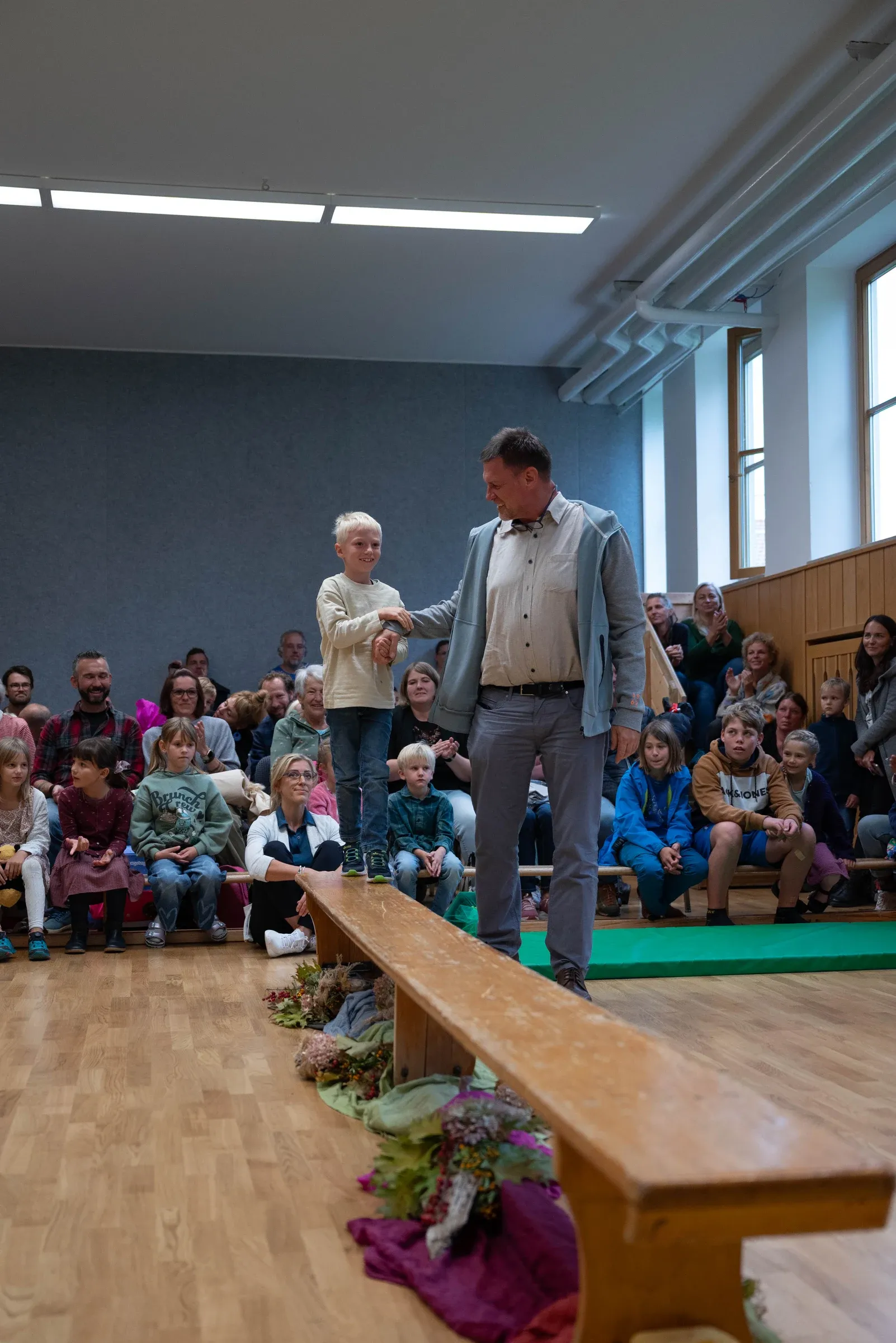 Ein Kind balanciert auf einer Holzbank und wird von einer zugewandten erwachsenen Begleitperson unterstützt